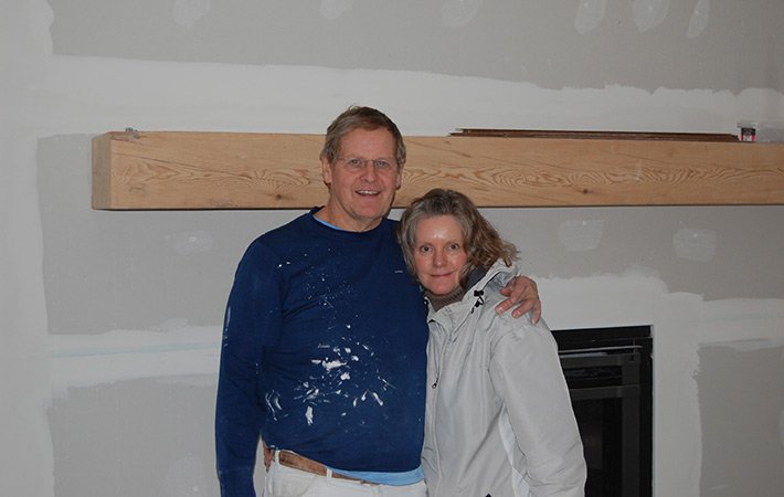 A man and woman stand together, smiling, in front of an unfinished wall with drywall patches. The man is wearing a blue shirt with white paint stains, and the woman is in a light-colored jacket.