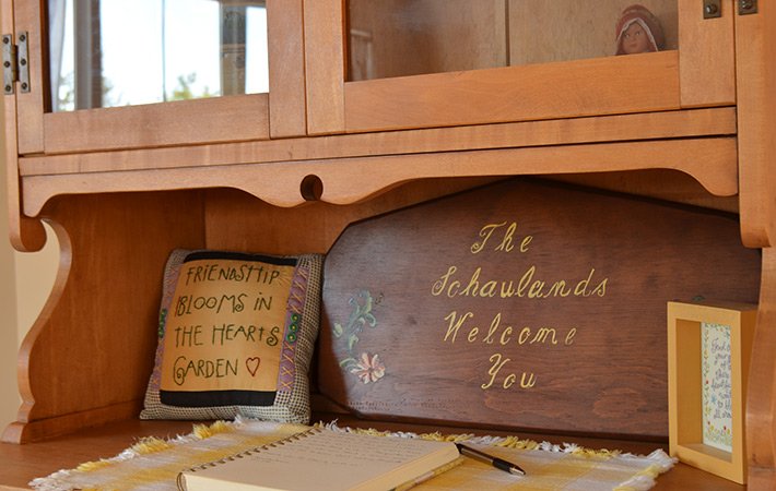 A wooden cabinet with a sign reading “The Schaulands Welcome You,” a pillow embroidered with “Friendship blooms in the heart’s garden,” a framed note, and an open notebook on a yellow-checked cloth.