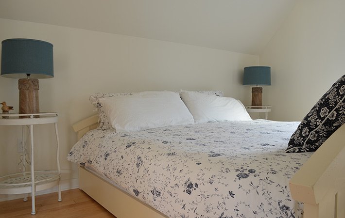 A neatly made bed with white and blue floral bedding, two white pillows, and a patterned black pillow. There are two round white bedside tables with blue lamps on each side, all set against cream-colored walls.