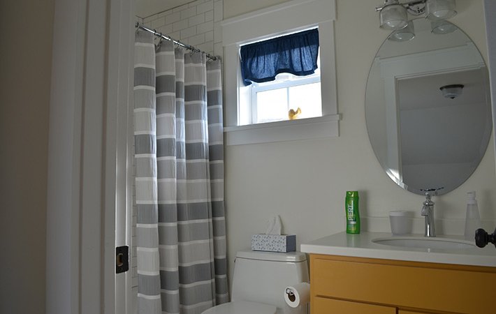 A bathroom with a striped gray and white shower curtain, a small window with a blue curtain, a box of tissues on the toilet, a yellow vanity, and a green spray can on the sink.