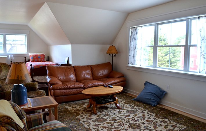 A cozy living room with a brown leather sofa, round wooden coffee table, lamp, and pillow on a patterned rug. A bed is seen in the back corner near a window, and large windows let in natural light.