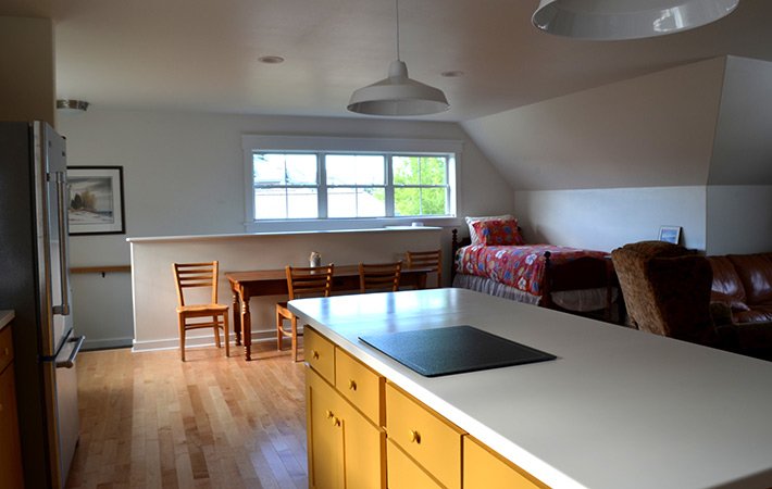 A bright, open-plan studio features a kitchen island with yellow cabinets, a dining table with chairs, a bed with a colorful quilt, a brown leather couch, and large windows letting in natural light.