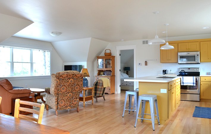 Bright open-plan living area with yellow kitchen cabinets, stainless steel appliances, two metal bar stools, upholstered armchairs, wooden furniture, and large window letting in natural light.