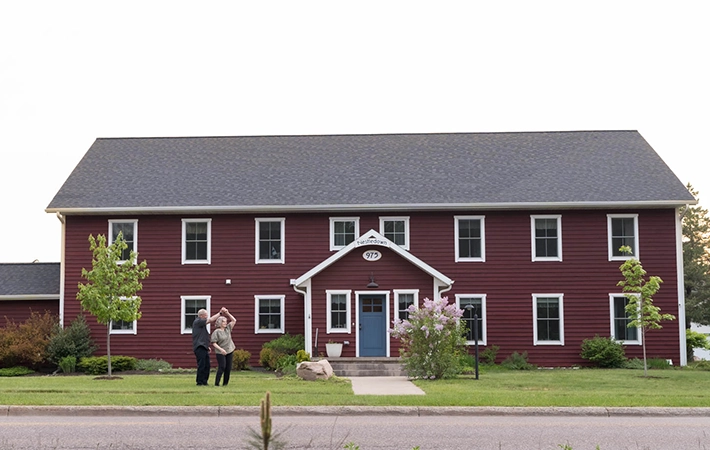 A large, two-story red house with white trim stands behind a neatly kept lawn. Two people stand talking near the walkway that leads to the front door, surrounded by small trees and shrubs.