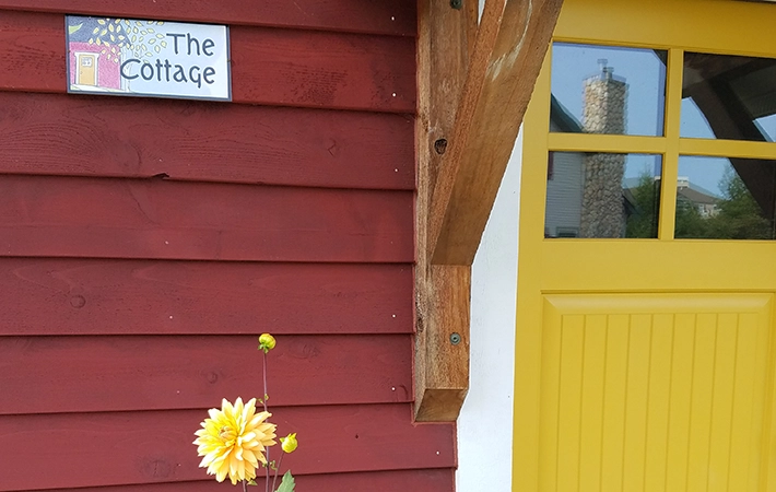 A red wooden building with a sign reading "The Cottage," a yellow door with window panes, and a yellow dahlia flower blooming in front.