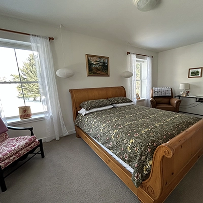 Bright bedroom with a wooden sleigh bed, floral bedspread, armchair, desk, and two windows with white curtains letting in natural light. A framed painting hangs above the bed.