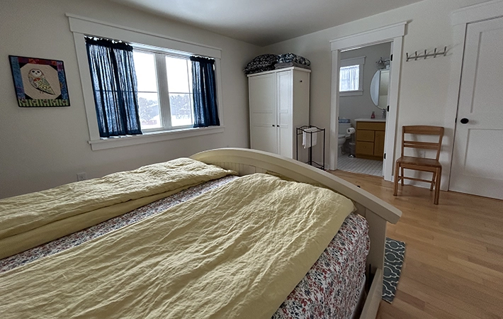 A bright bedroom with a bed covered in yellow bedding, a window with dark blue curtains, a white wardrobe, a wooden chair, and an open door leading to a bathroom. A colorful owl picture hangs on the wall.