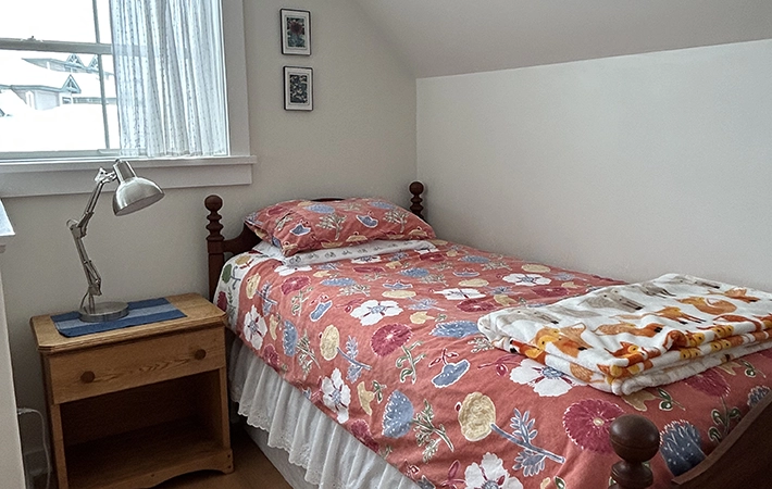 A small, tidy bedroom with a single bed covered in a floral bedspread, a folded blanket at the foot, a wooden nightstand with a lamp, and a window with sheer curtains letting in natural light.