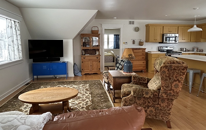 A cozy living room with patterned armchairs, a brown sofa, wooden coffee table, TV on a blue stand, rug, and a view into a kitchen with yellow cabinets and stainless steel appliances. Natural light enters through a window.