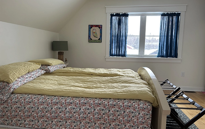 A cozy bedroom with a bed covered in floral-patterned bedding and yellow blankets, a green lamp on a bedside table, a framed owl picture on the wall, blue curtains, and a window letting in natural light.