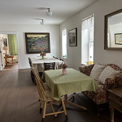 A cozy, well-lit dining area with two tables, chairs, a floral loveseat, artwork on the walls, large mirrors, and windows letting in natural light. The room extends into a living space with a lamp and green curtains.