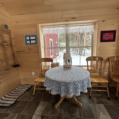 A cozy wooden room with a round table covered by a blue patterned cloth, surrounded by four chairs, and a window overlooking a snowy yard. Finnish and Norwegian flags hang on the walls.